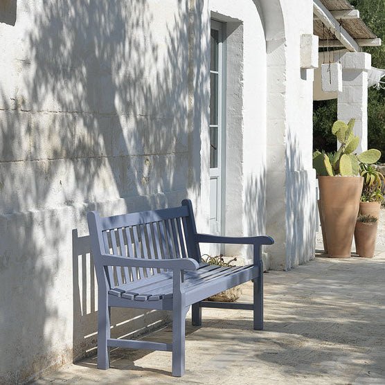 Blue wooden outdoor bench with a traditional slatted design, placed against a white Mediterranean-style home with soft shadows from olive trees. | Ethimo Notting Hill