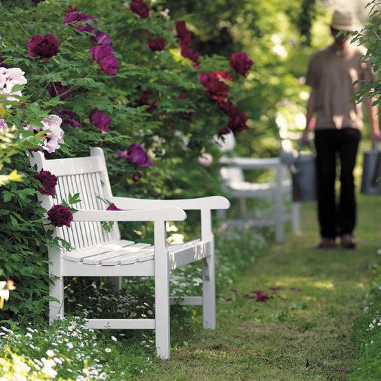 Compact white wooden bench with armrests, placed along a floral pathway with blooming red and purple flowers. | Ethimo Notting Hill