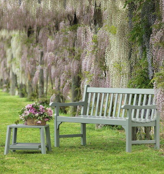 Green wooden garden bench with matching side table, placed under cascading wisteria flowers in a peaceful outdoor setting. | Ethimo Notting Hill