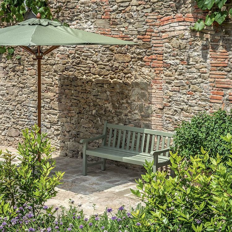 Green wooden outdoor bench with classic slatted backrest, placed against a rustic stone wall with a green umbrella providing shade. | Ethimo Notting Hill