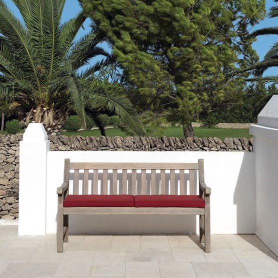 Pickled teak wooden bench with a slatted backrest, featuring deep red seat cushions, placed on a white stone patio with greenery in the background. | Ethimo Notting Hill