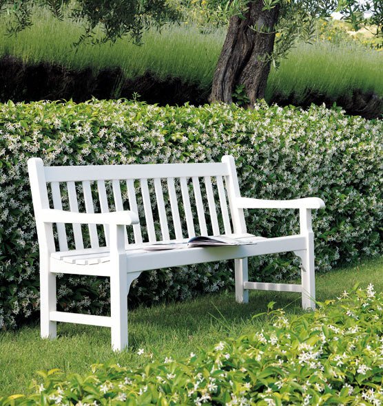  White wooden garden bench with a classic slatted design, positioned in a lush green garden surrounded by flowering hedges. | Ethimo Notting Hill